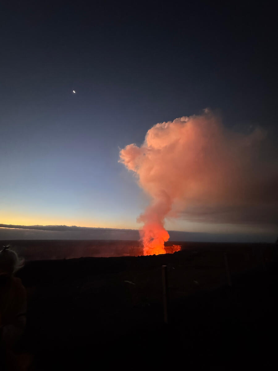 Photo of Joan Obra in a flowered dress, sitting atop a stone wall at a beach on Hawai'i Island.